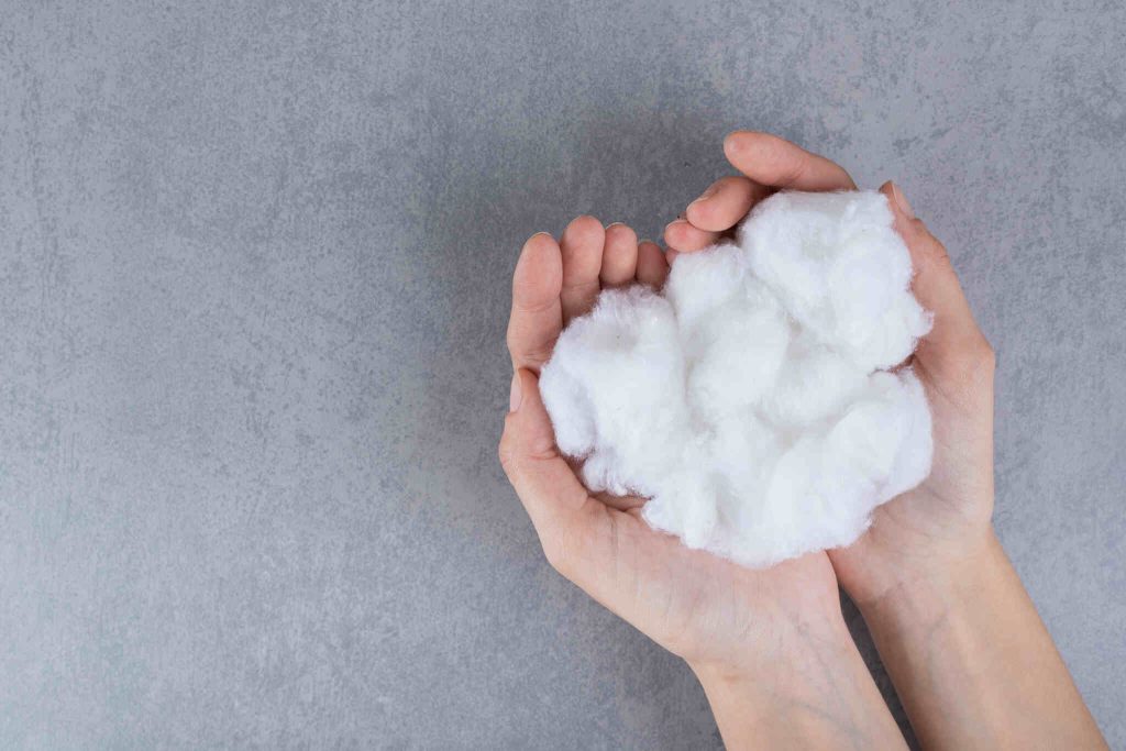Close de duas mãos abertas segurando um punhado generoso de algodão hidrofílico branco e macio. O algodão tem uma textura fibrosa e volumosa, contrastando com o fundo de textura cinza neutra que imita cimento queimado.