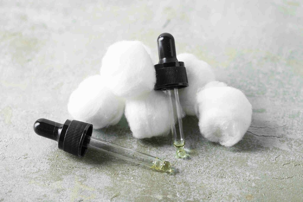 Pequenas bolas de algodão branco agrupadas ao lado de dois conta-gotas de vidro com tampas pretas. Um dos conta-gotas está deitado, mostrando um líquido levemente amarelado na ponta. O cenário sugere cuidados com a pele ou procedimentos estéticos sobre uma superfície rústica cinza.