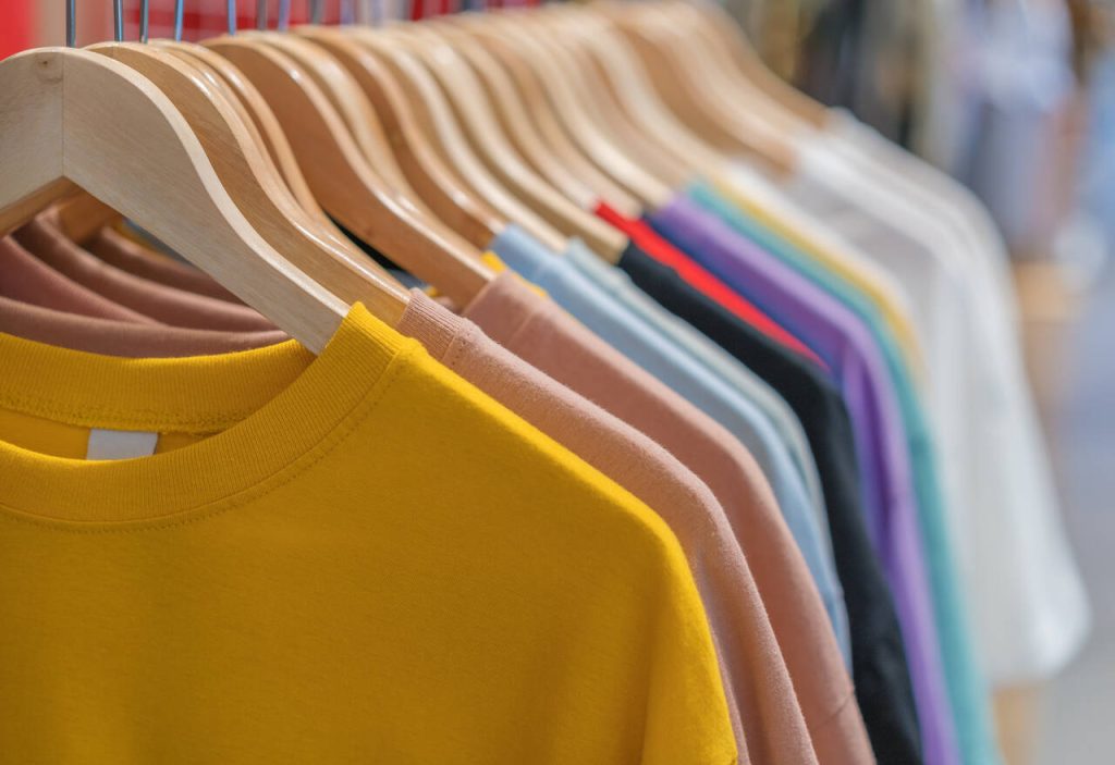 Fileira de camisetas de algodão coloridas penduradas em cabides de madeira em uma loja de roupas, destacando diferentes tons de malha.