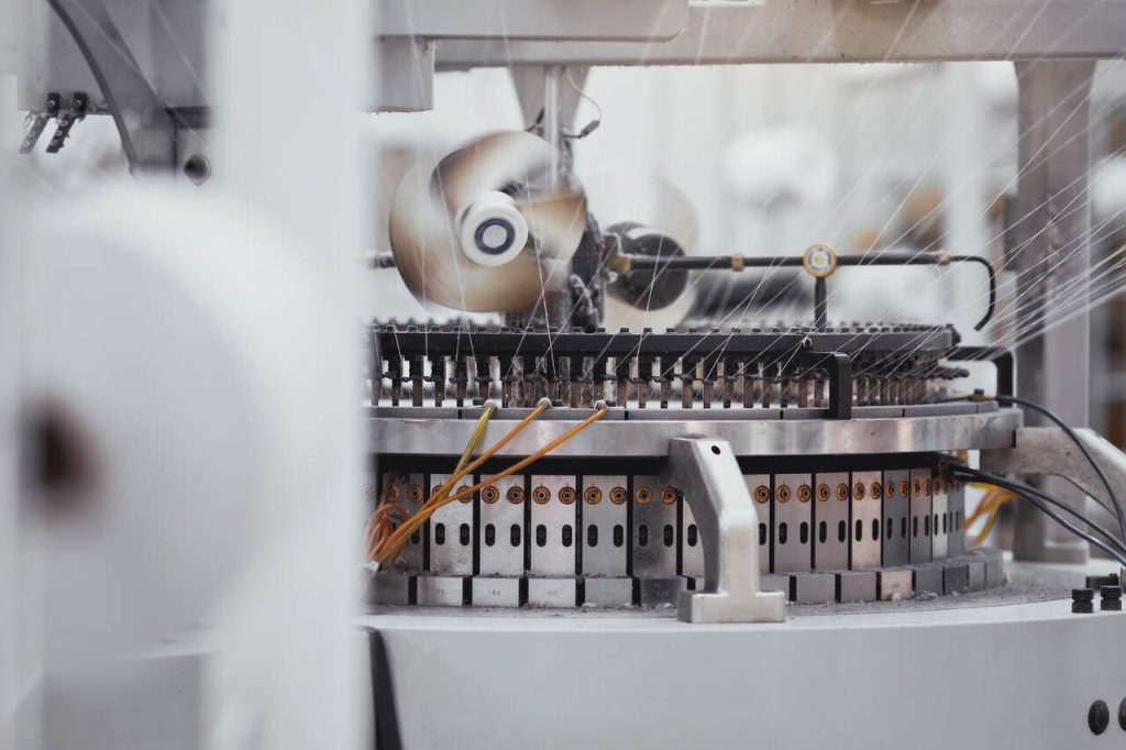 Máquina de tecelagem circular industrial em operação em uma fábrica têxtil, processando fios de algodão branco para produção de tecidos.