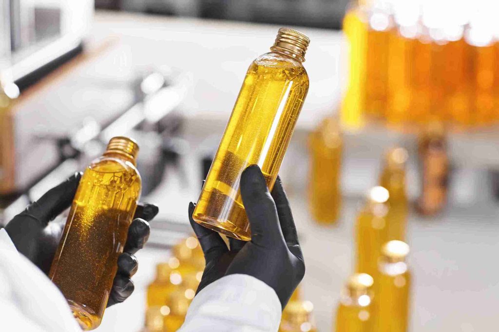 Mãos com luvas pretas segurando frascos de óleo de algodão dourado e límpido em ambiente laboratorial ou industrial.