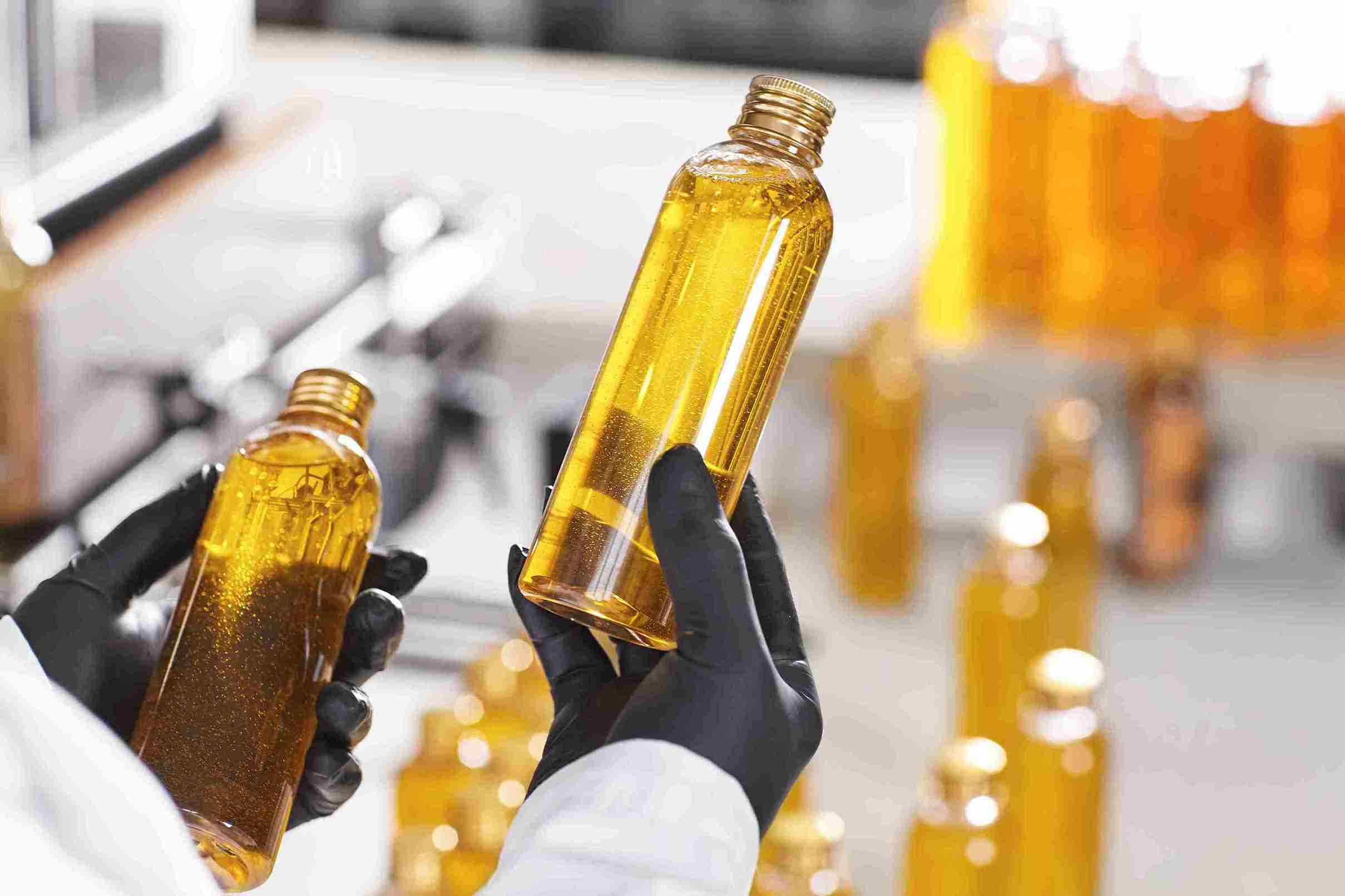 Mãos com luvas pretas segurando frascos de óleo de algodão dourado e límpido em ambiente laboratorial ou industrial.