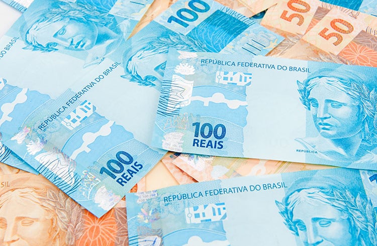 Diversas cédulas de 100 e 50 Reais espalhadas de forma desordenada sobre uma superfície clara. Em destaque, as notas azuis de 100 Reais exibem a efígie da República e a inscrição "República Federativa do Brasil". Ao fundo e nas laterais, partes das notas de 50 Reais em tons de laranja também são visíveis.
