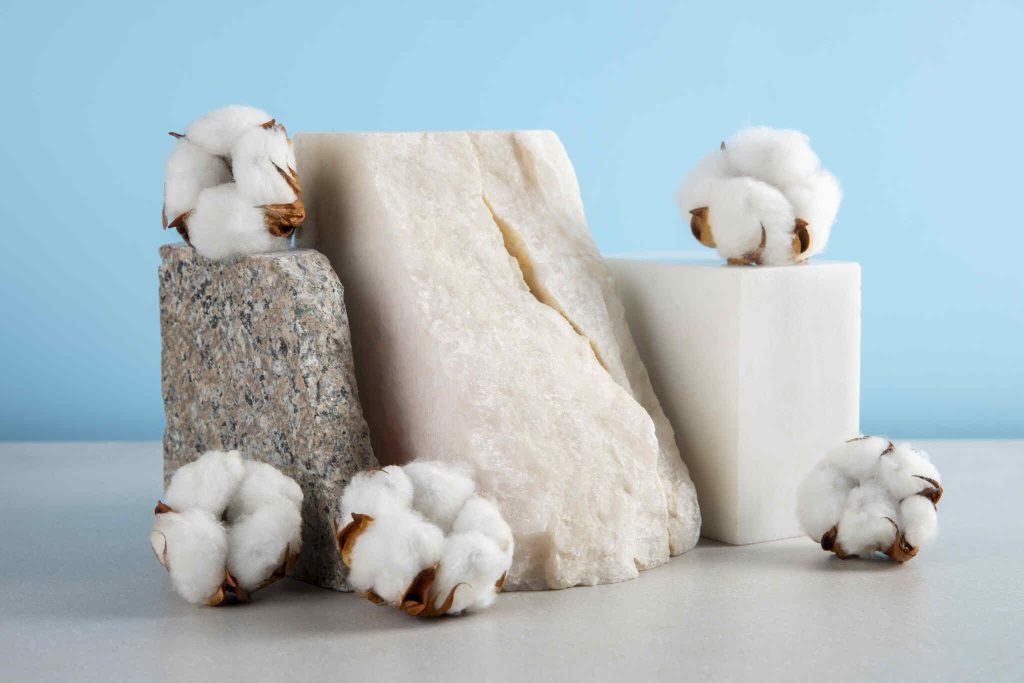 Capulhos de algodão branco natural posicionados sobre blocos de pedra e superfícies geométricas brancas em um fundo azul claro.