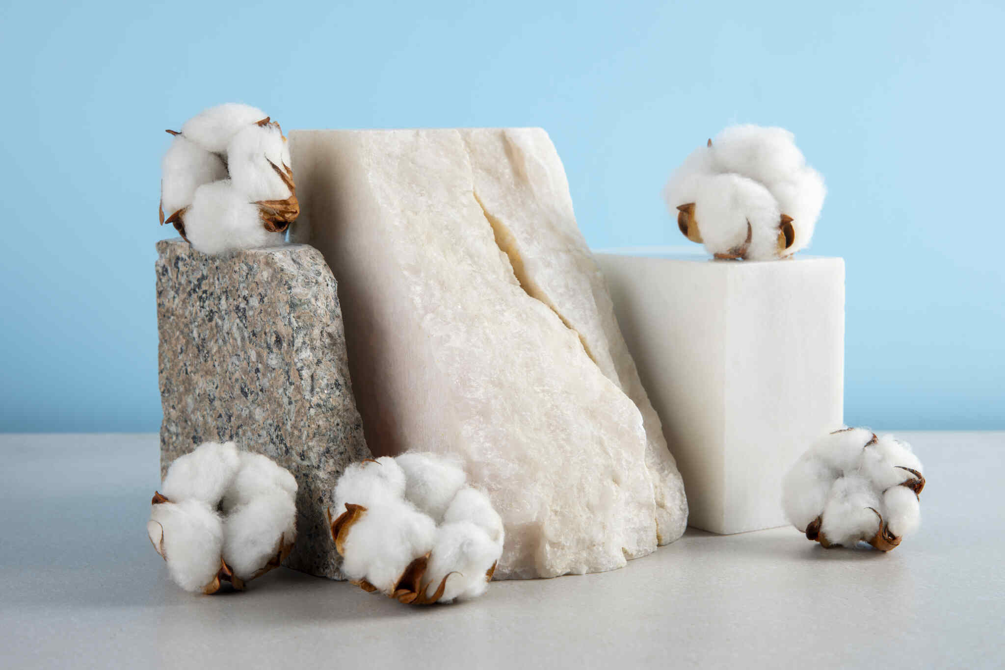 Capulhos de algodão branco natural posicionados sobre blocos de pedra e superfícies geométricas brancas em um fundo azul claro.